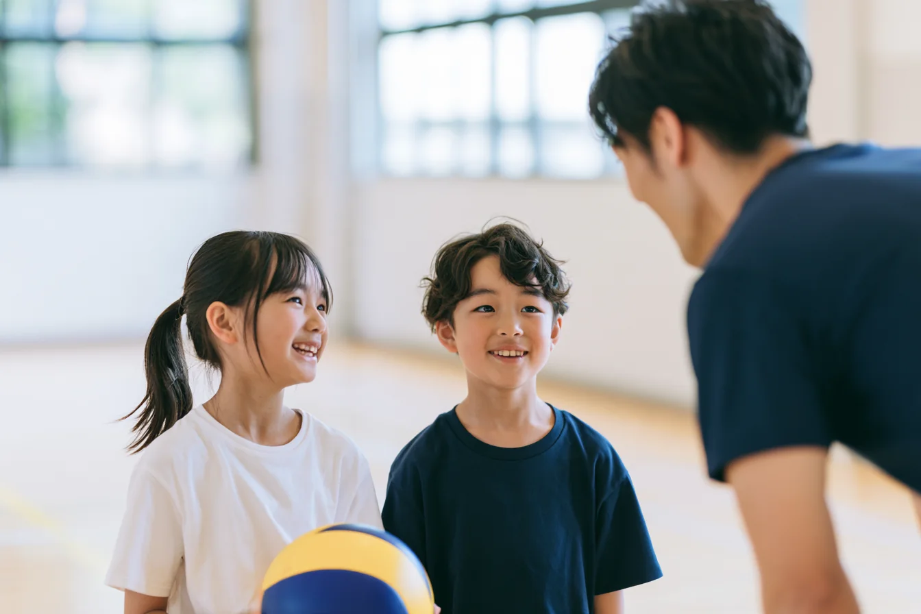 体育館でコーチと話す、バレーボールを持った子どもたち