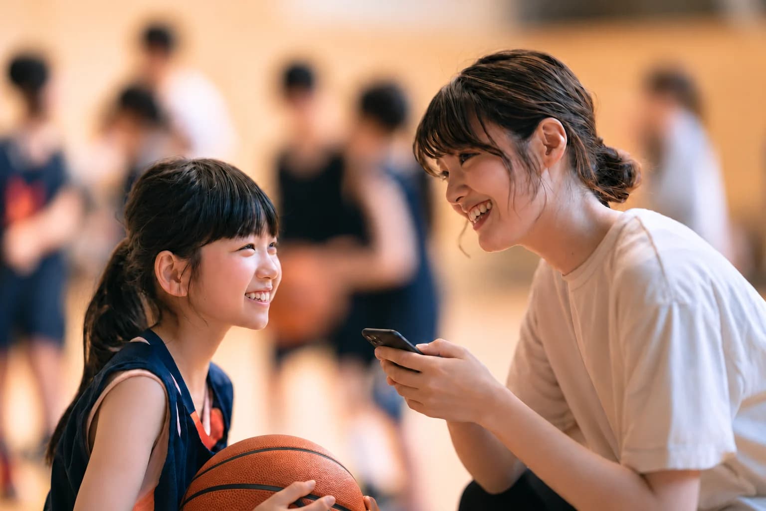 体育館で、スマートフォンを手にした保護者と、バスケットボールを抱えた子どもが笑顔で話している様子